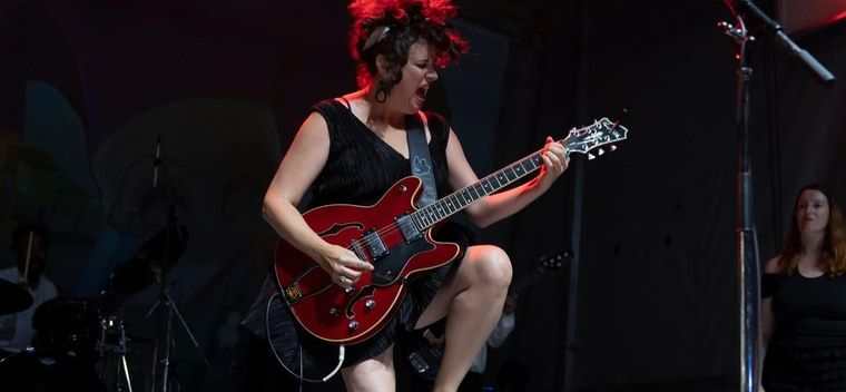 A guitarist mid song on a dark stage, lit red, playing a red hollow body guitar